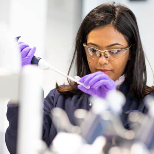 Woman in lab coat, gloves and goggles holding a pippette